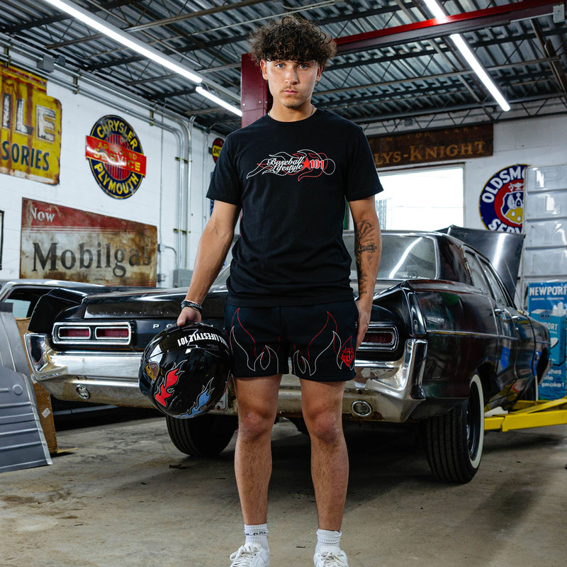 Young man standing in vintage garage holding racing helmet, wearing black Flames Stitched tee and matching black flame sweatshorts, classic cars in background.