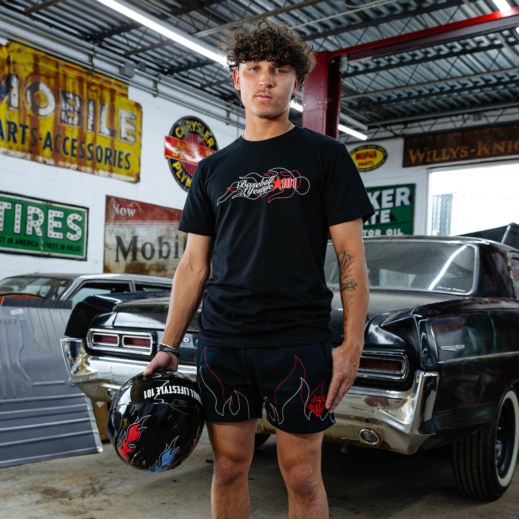 Young man standing in vintage garage holding racing helmet, wearing black Flames Stitched tee with BL101 flame graphic and black flame shorts, classic cars in background.