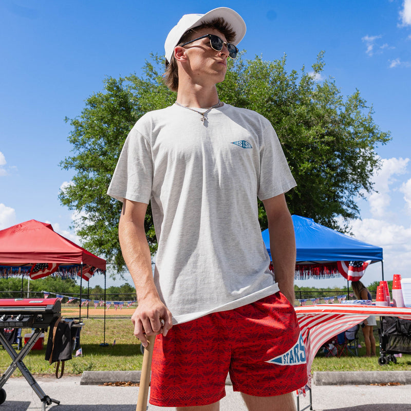 Full body adult male model wearing red Freedom Swings shorts with heather gray tee and white snapback holding baseball bat at outdoor baseball event