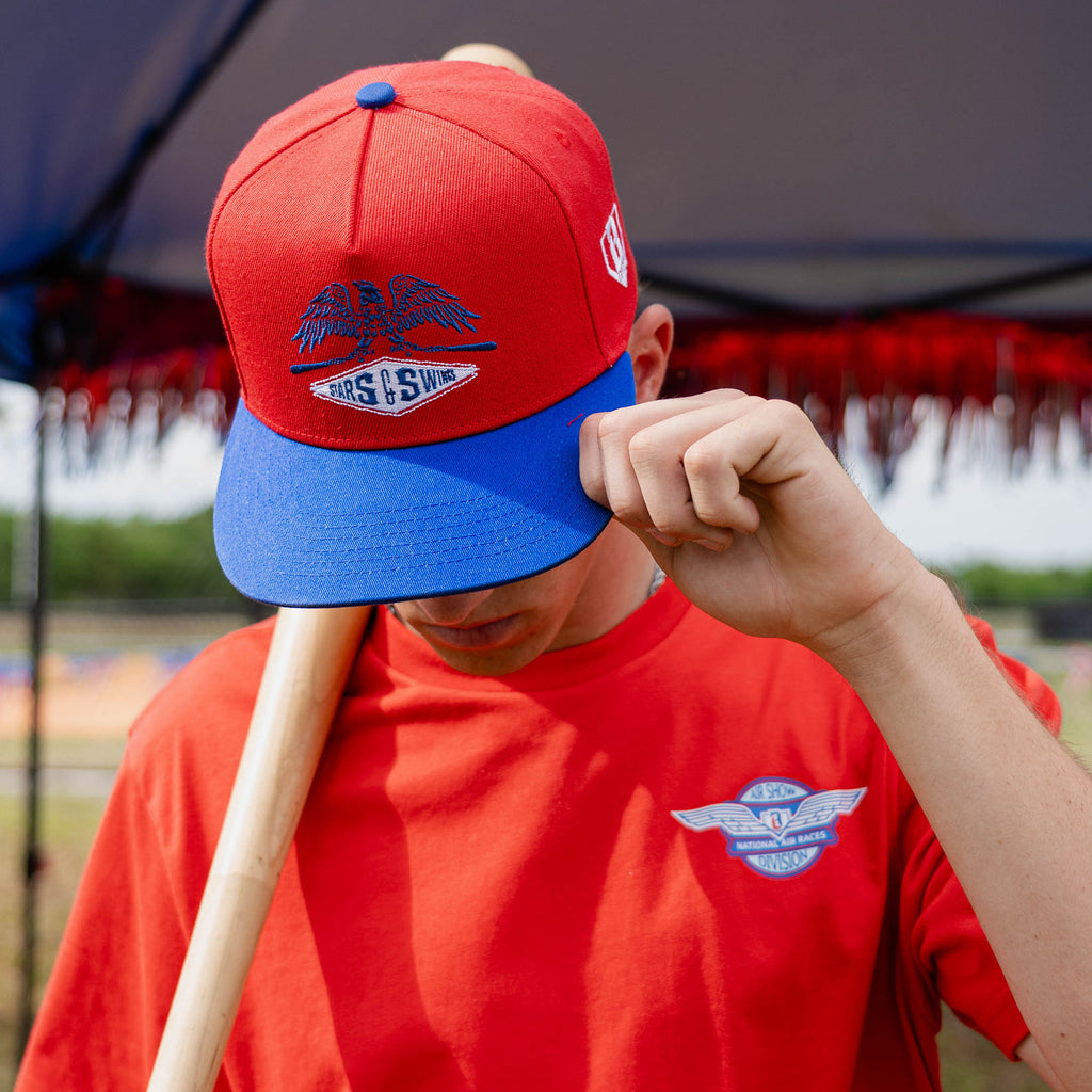 Person wearing a red and blue cap with a Freedom Swings logo, holding a baseball bat, against a blurred outdoor background.