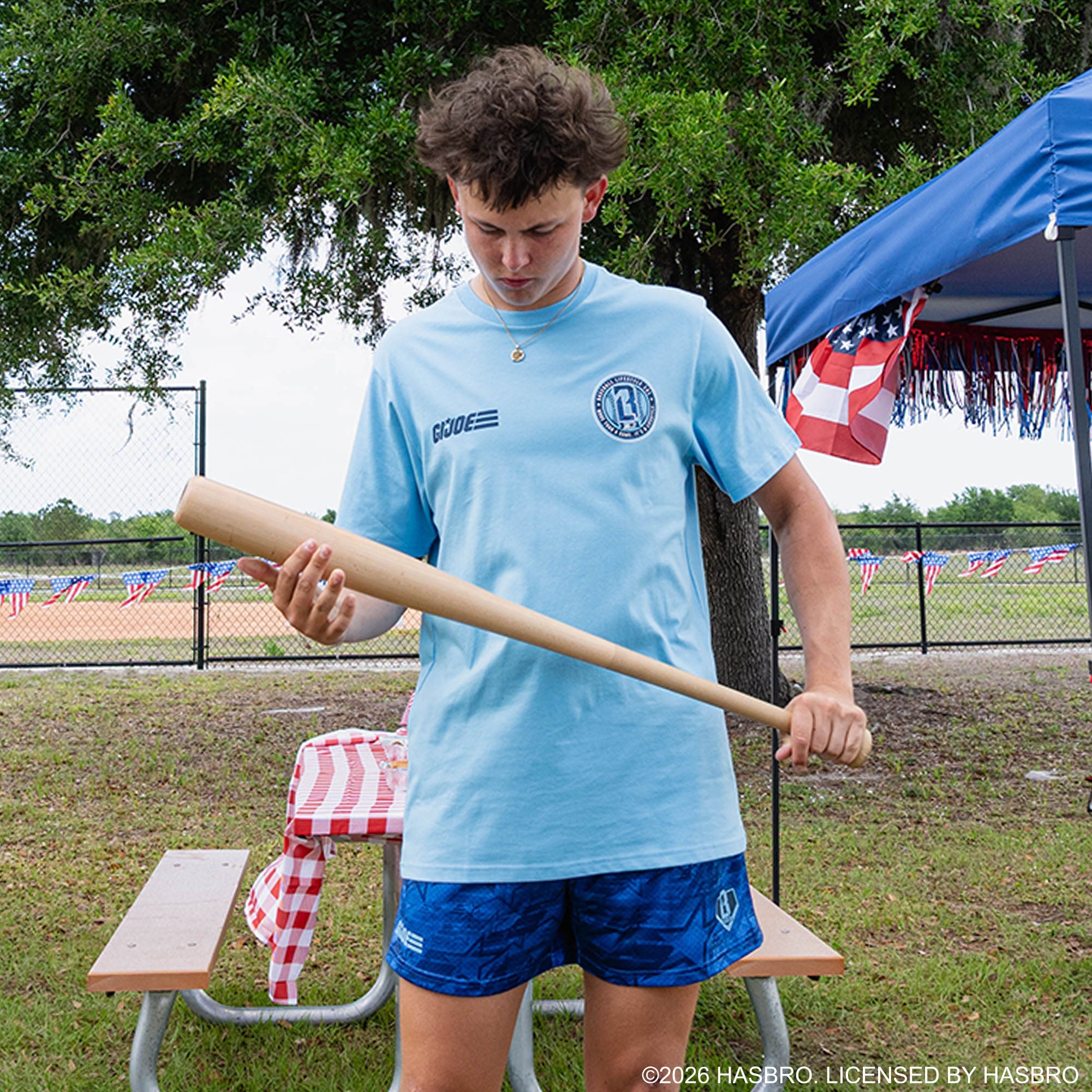 Adult male model wearing light blue GI Joe tee examining baseball bat at outdoor baseball event