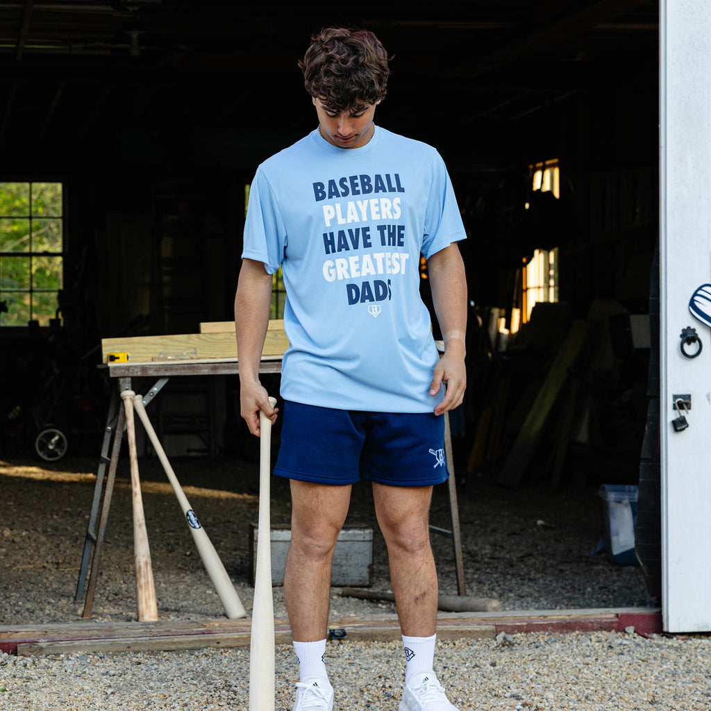 Young adult male model looking down while holding a wooden baseball bat and wearing the navy Heritage Diamond Sweatshort paired with the columbia blue Greatest Dad Athletic Tee and white BL101 crew socks.