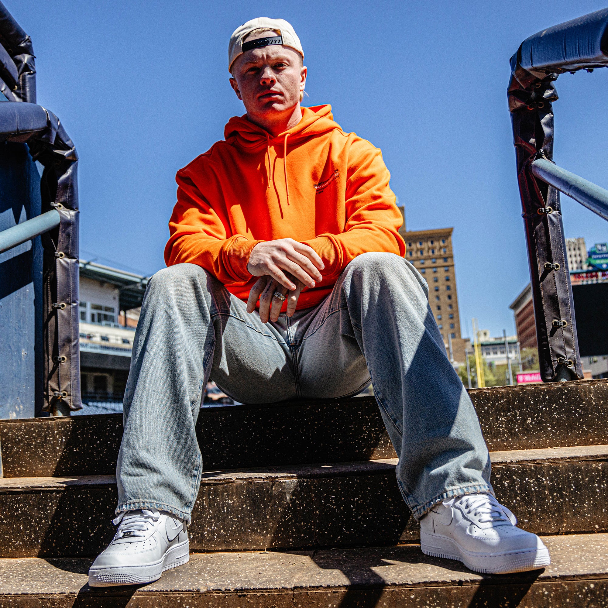 Person wearing an orange hoodie and light blue jeans sitting on steps with a cityscape background