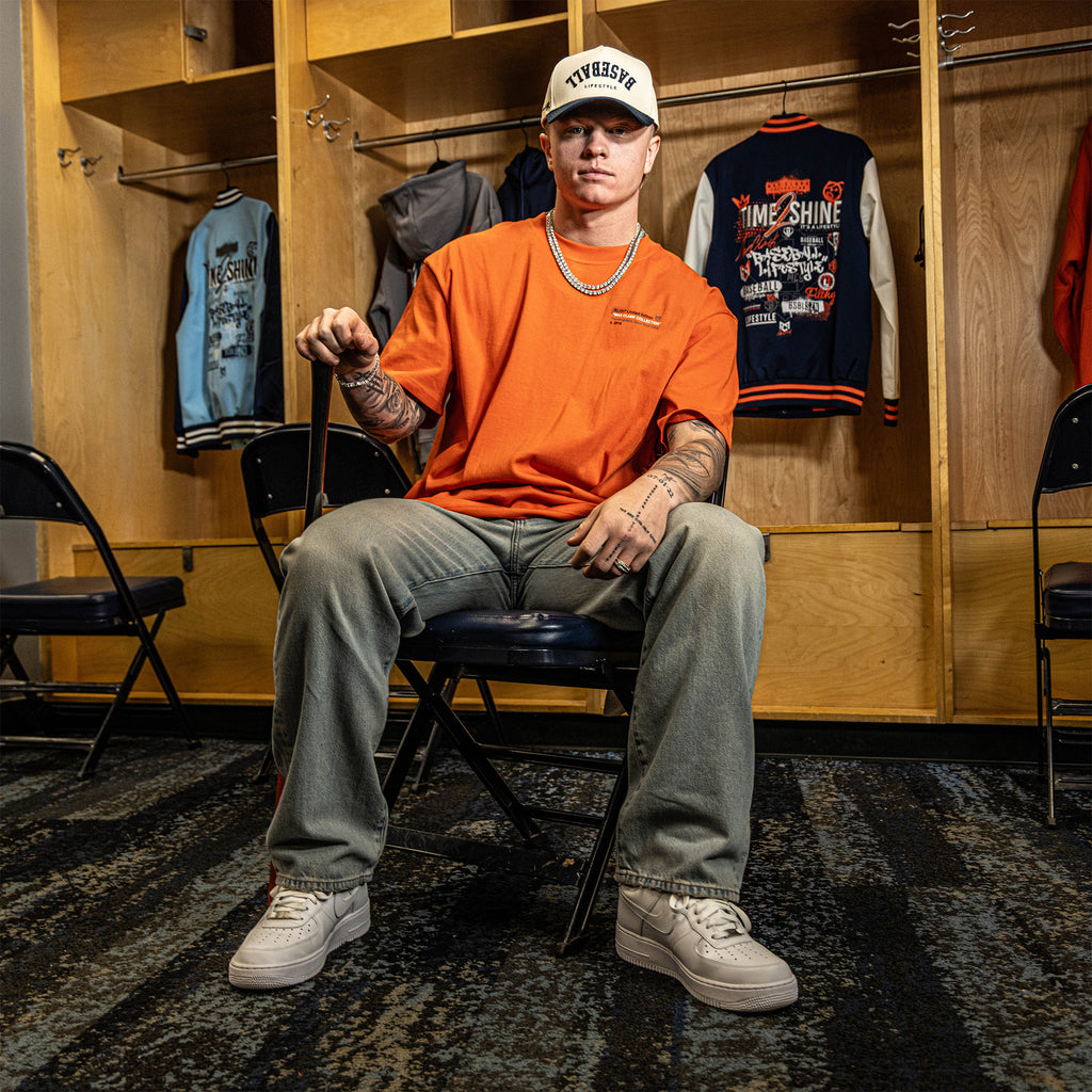Person sitting in a locker room wearing an orange shirt and gray pants.