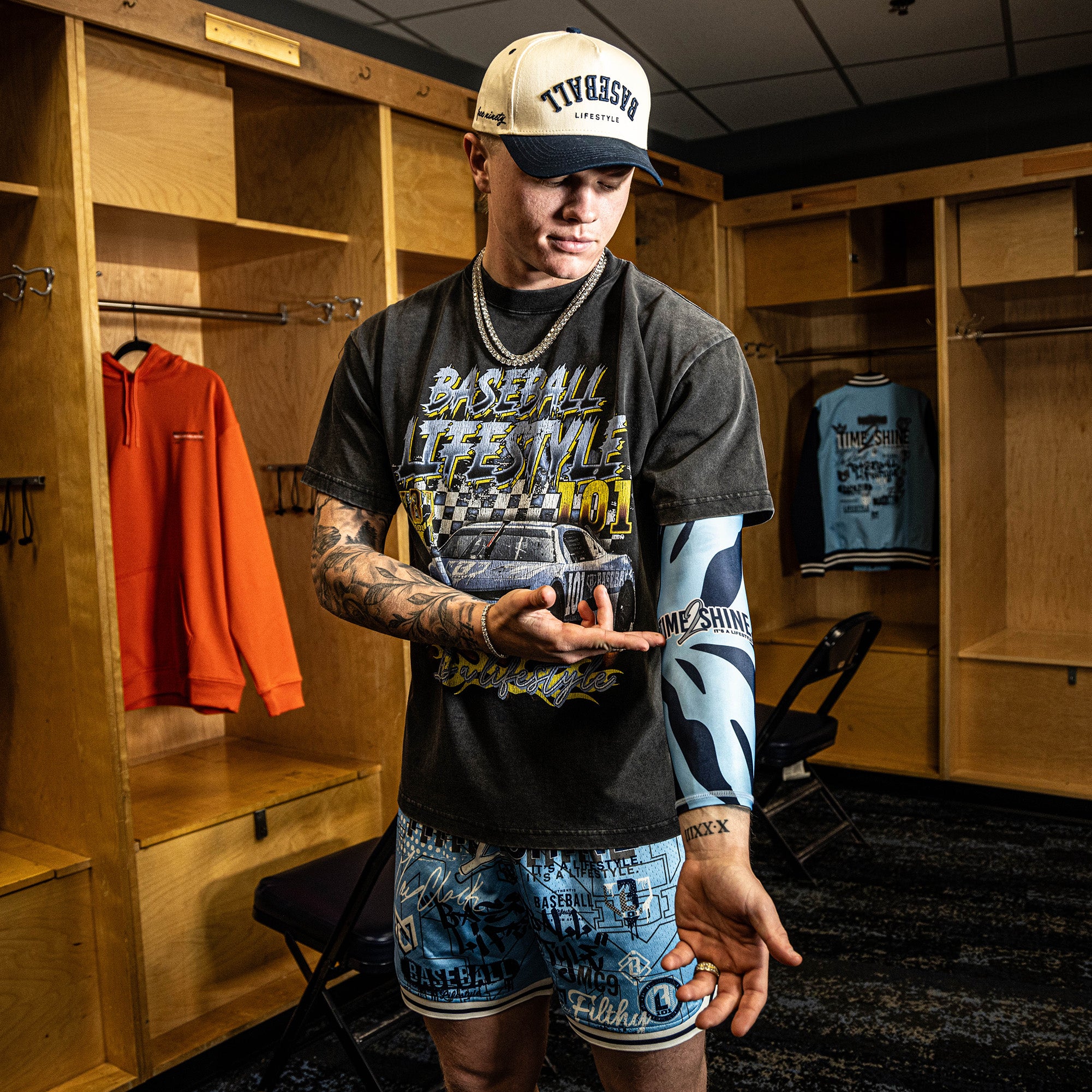 Man in a locker room wearing a graphic t-shirt and shorts, holding a baseball.