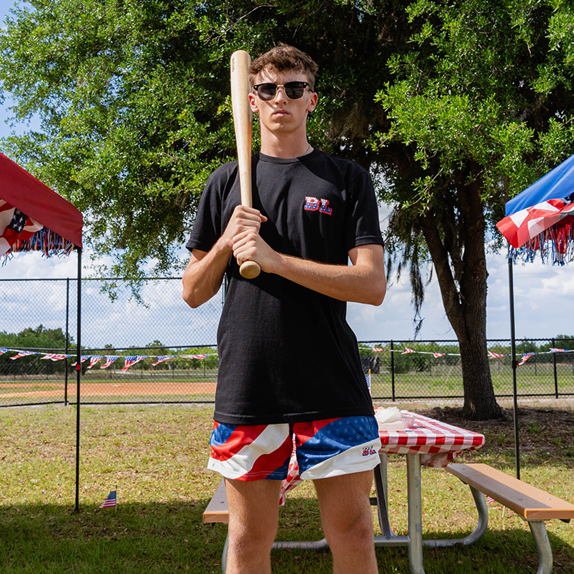 Full body adult male model wearing red white and blue Patriot Pitch shorts with black tee holding baseball bat at outdoor baseball event