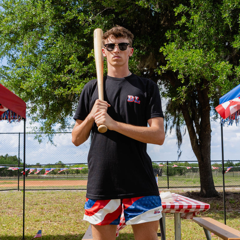 Front-facing adult male model holding baseball bat wearing black Patriot Pitch tee with red white and blue shorts at outdoor baseball event