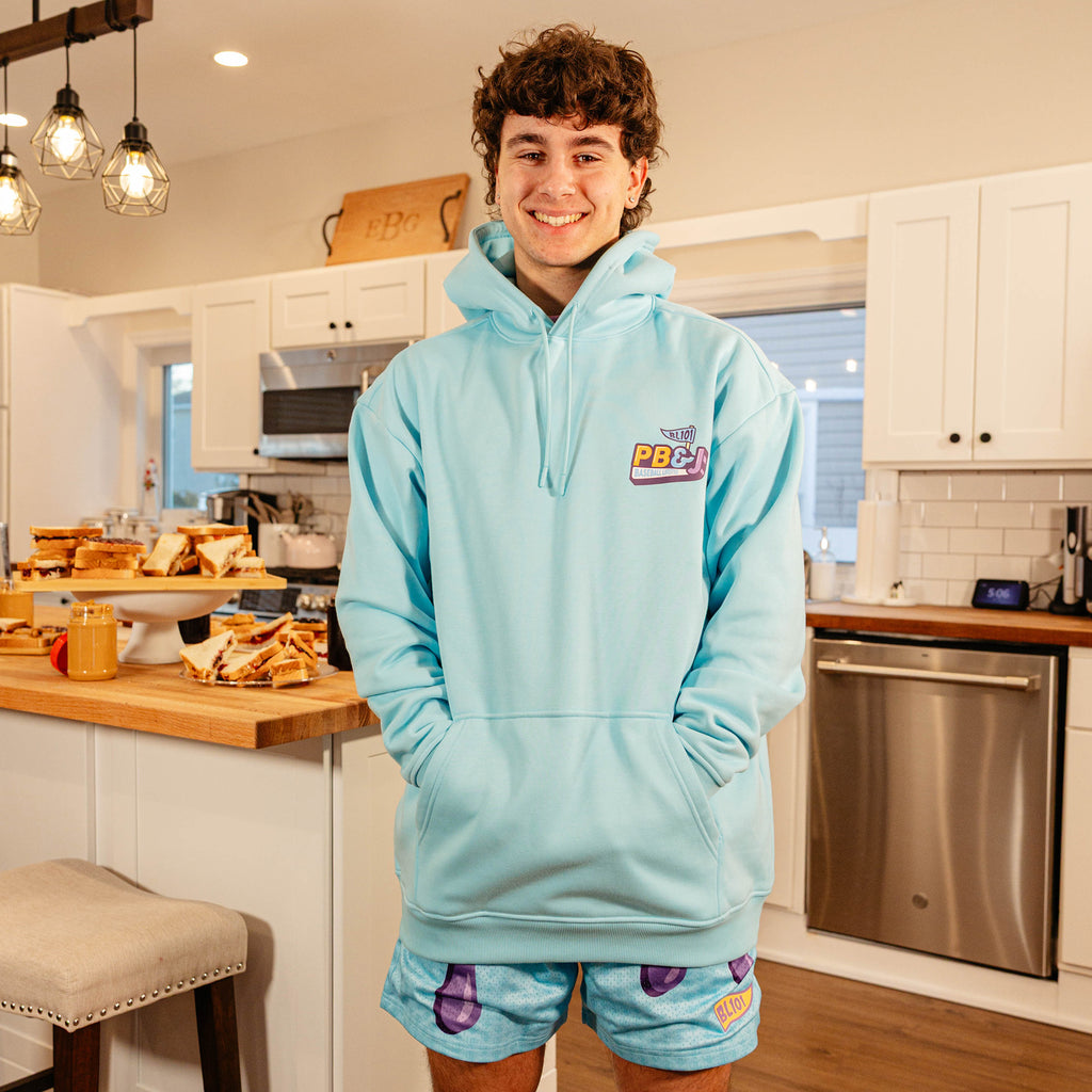 Male model smiling in a kitchen wearing the BL101 PB&J Baseball Lifestyle light blue hoodie with matching PB&J mesh shorts.