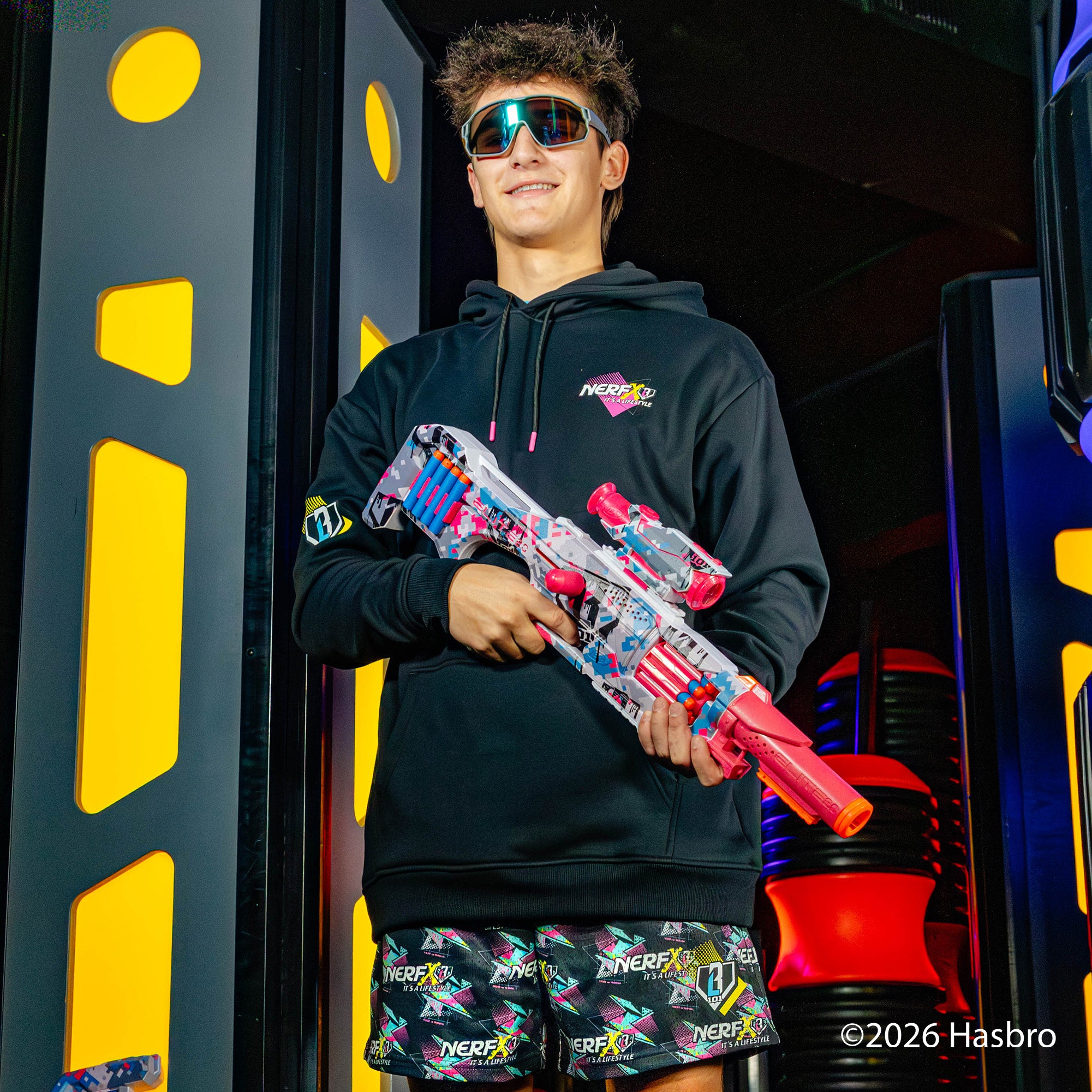 Person wearing a black hoodie and colorful shorts holding a Nerf gun in front of a colorful background.