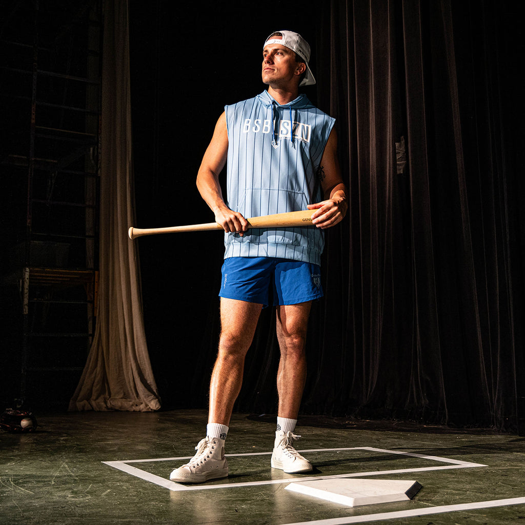 Adult male model holding a wood bat on a stage set wearing the blue Jackson Olson x BL101 Pro Series Shorts V2 paired with a matching light blue pinstripe sleeveless hoodie
