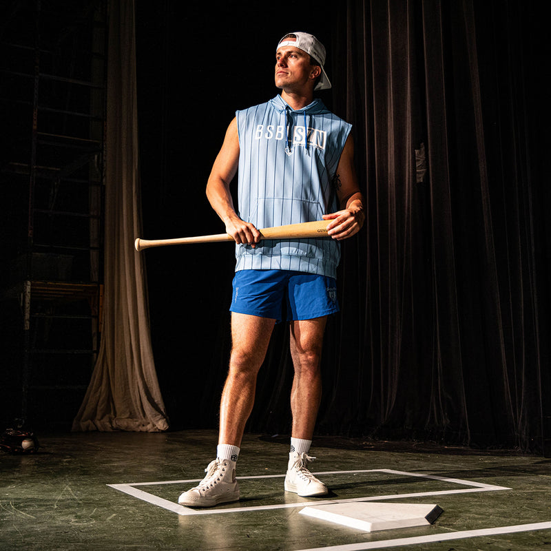 Adult male model holding a wood bat on a stage set wearing the blue Jackson Olson x BL101 Pro Series Shorts V2 paired with a matching light blue pinstripe sleeveless hoodie