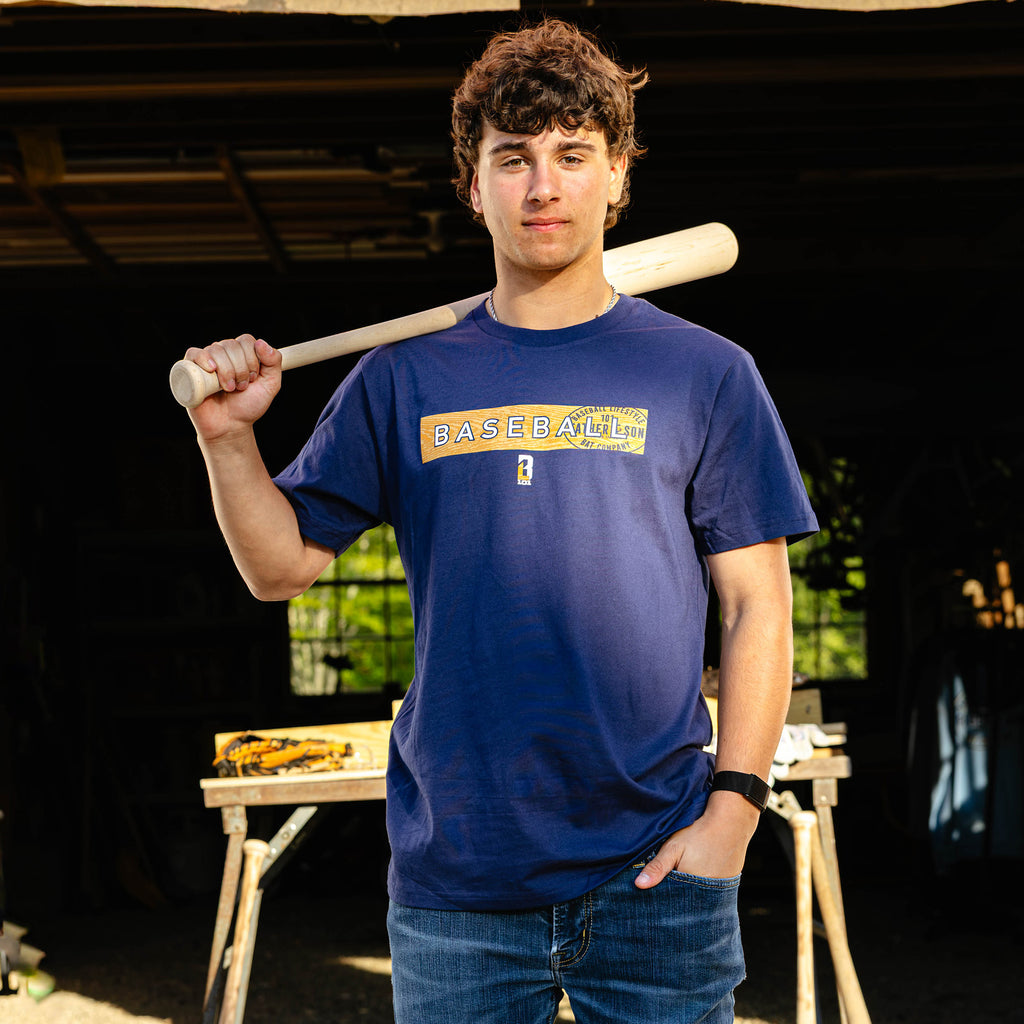 Young adult male model resting a wooden baseball bat on his shoulder while wearing the navy BL101 Father & Son Bat Company Radar Tee in a woodshop setting.