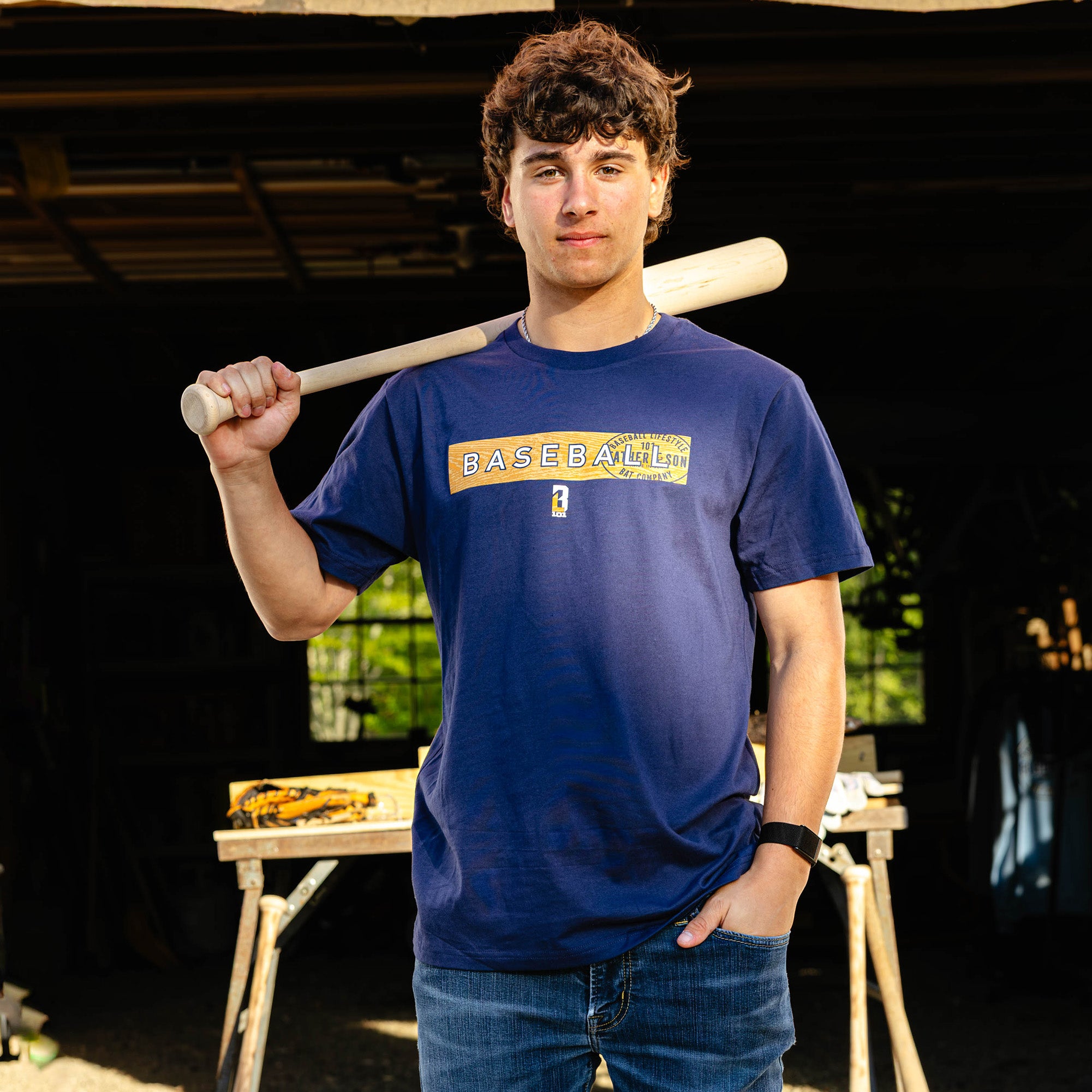 Young adult male model resting a wooden baseball bat on his shoulder while wearing the navy BL101 Father & Son Bat Company Radar Tee in a woodshop setting.