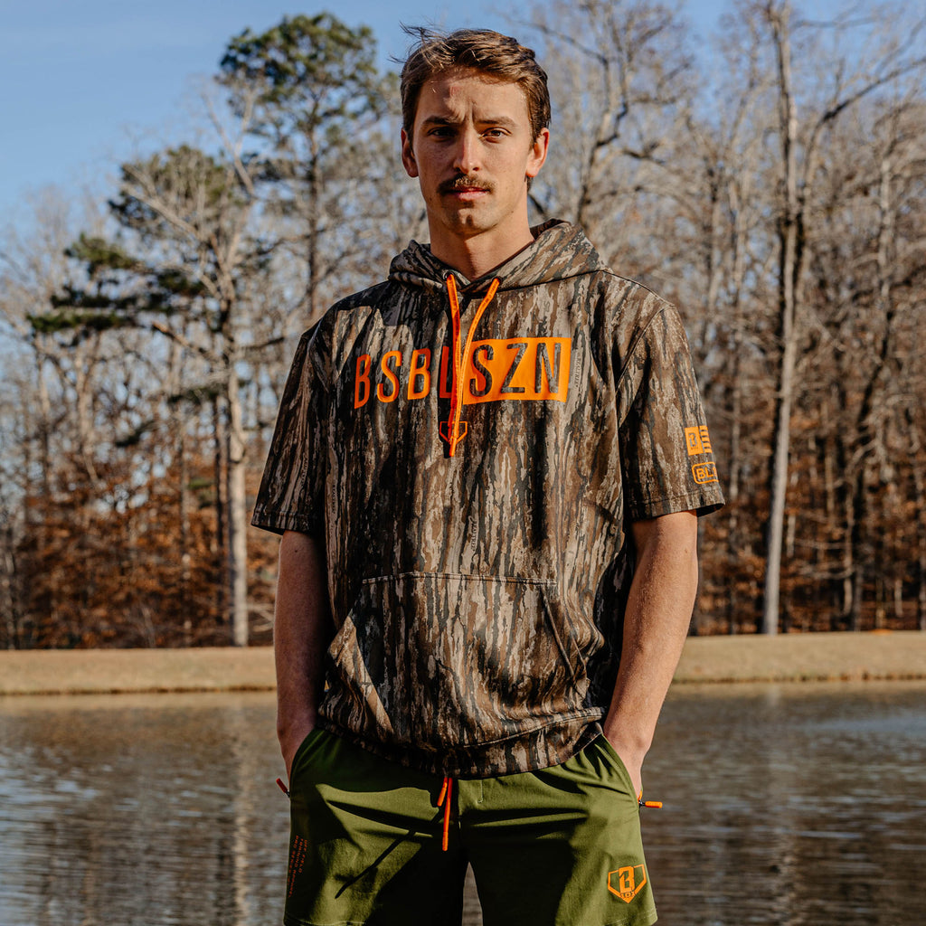 Man wearing a camouflage hoodie with orange 'BSBL SZN' text standing by a body of water with trees in the background.
