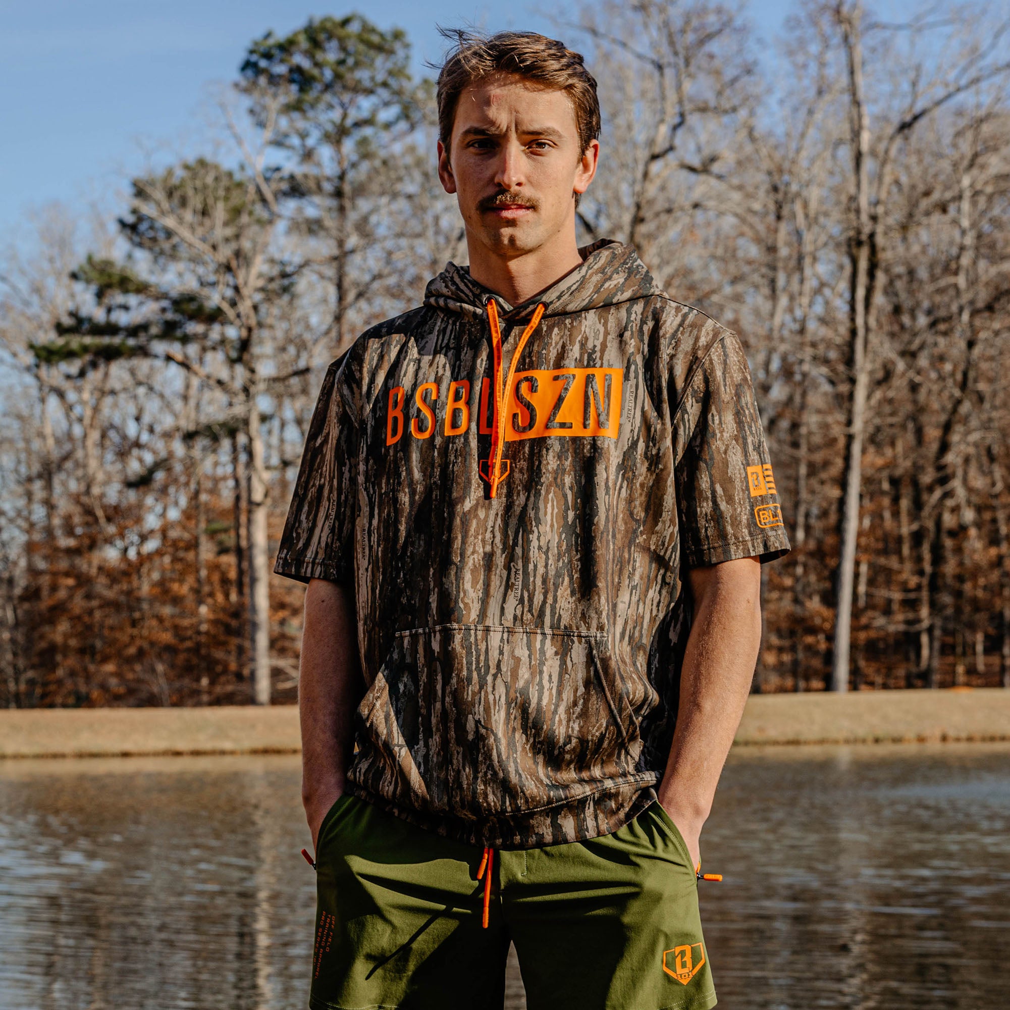 Man wearing a camouflage hoodie with orange 'BSBL SZN' text standing by a body of water with trees in the background.