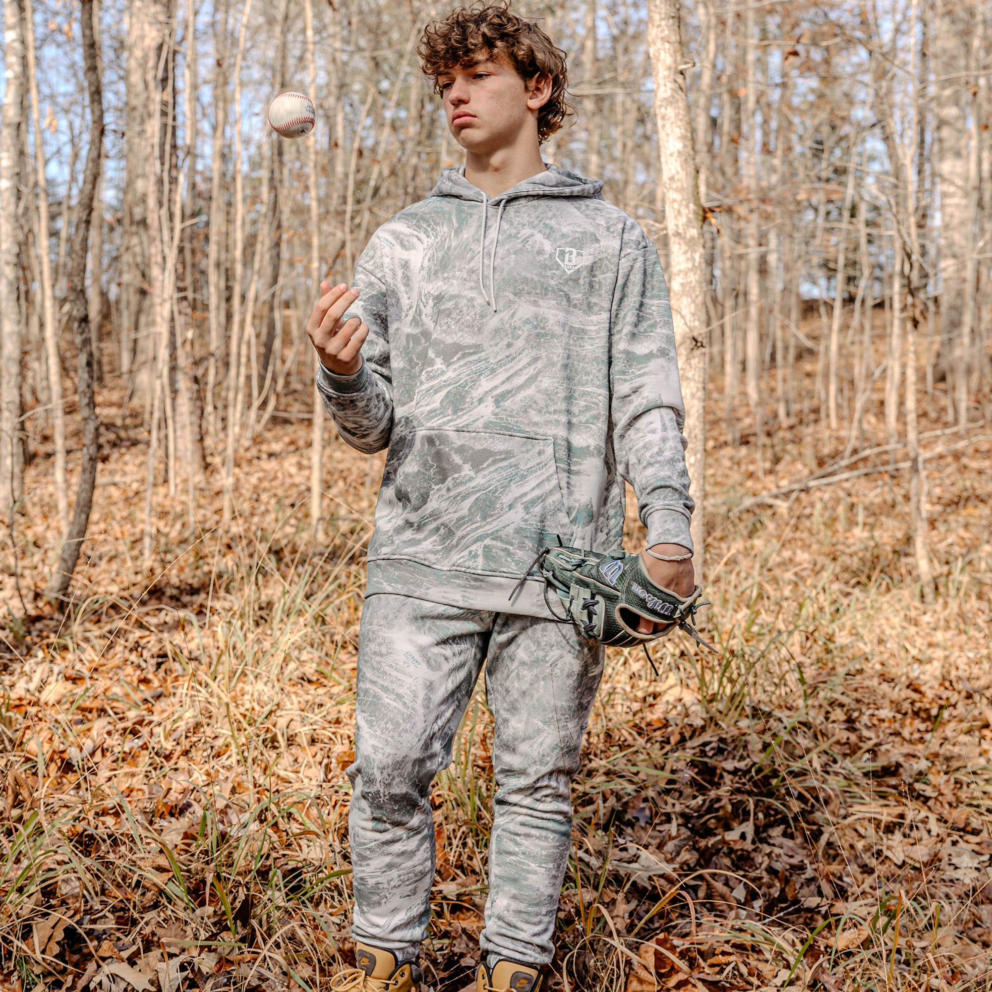 Person in light camouflage clothing holding a baseball and glove in a forest setting