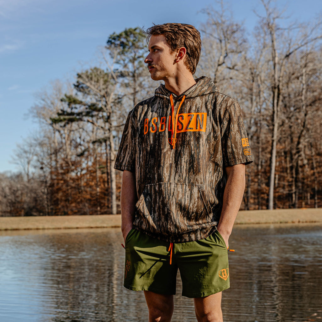 Person wearing a camouflage hoodie with orange 'BSBL SZN' text and green shorts standing by a body of water.