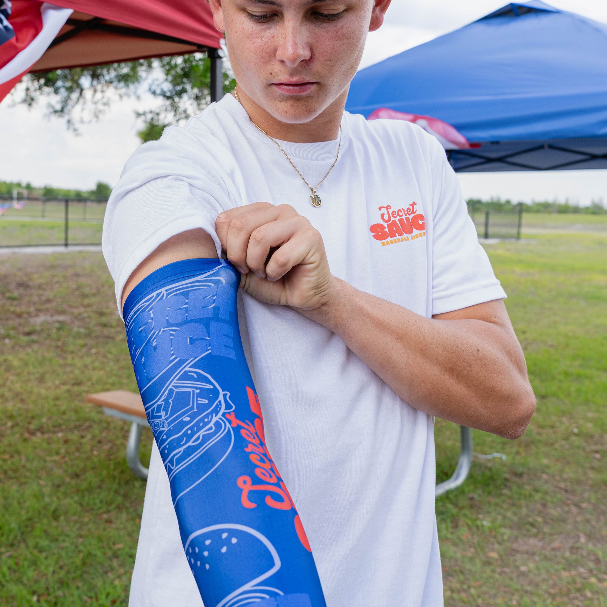 Adult male model flexing arm showing blue Secret Sauce arm sleeve with burger line art pattern and Secret Sauce logo while wearing white Secret Sauce tee at outdoor baseball event