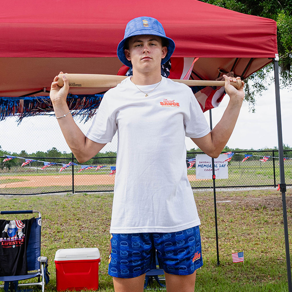 Person holding a baseball bat under a red tent wearing a white t-shirt with 'Secret Sauce' text
