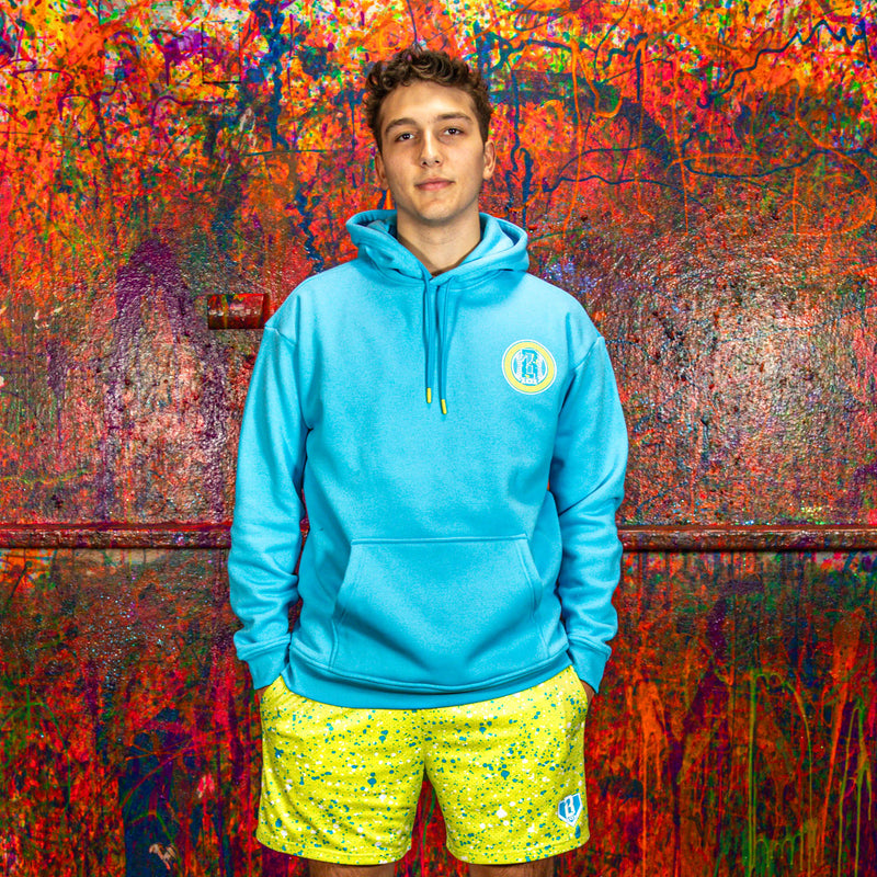 Person wearing a blue hoodie and yellow shorts standing against a colorful, textured wall.
