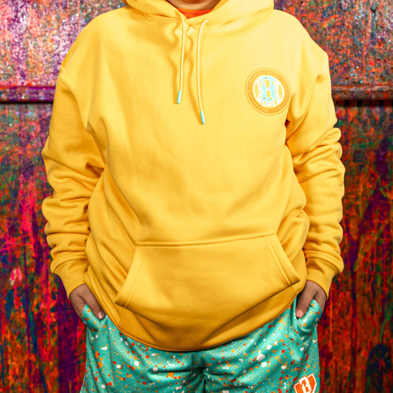 Person wearing a orange hoodie with a logo and teal shorts with a pattern, standing against a colorful abstract background.
