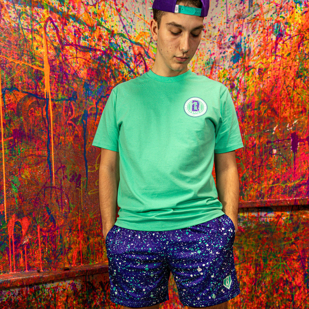 Person wearing a green t-shirt with a BL101 logo and purple shorts with a pattern, standing in front of a colorful graffiti wall.