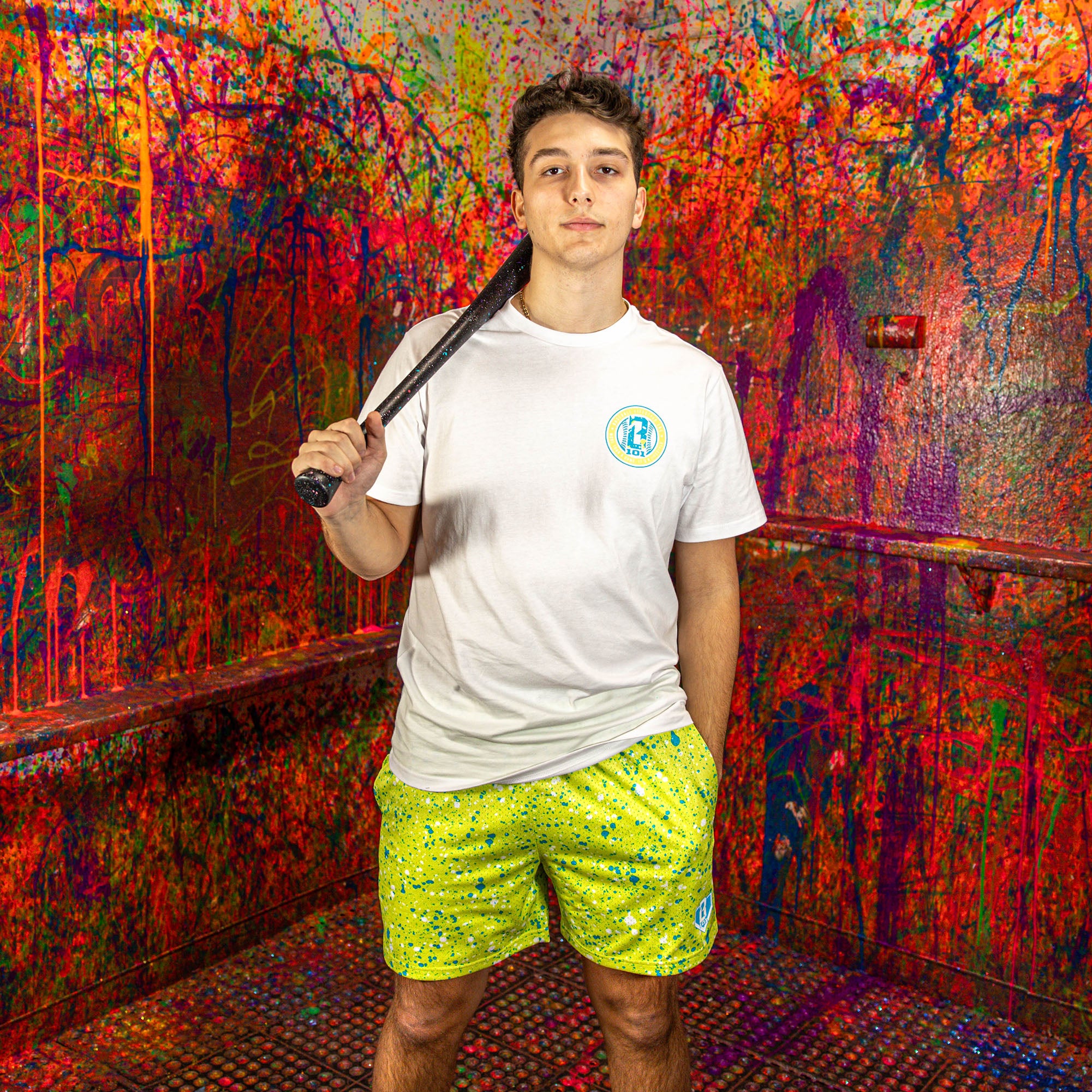 Person wearing a white shirt and yellow shorts while holding a baseball bat in front of a splattered wall