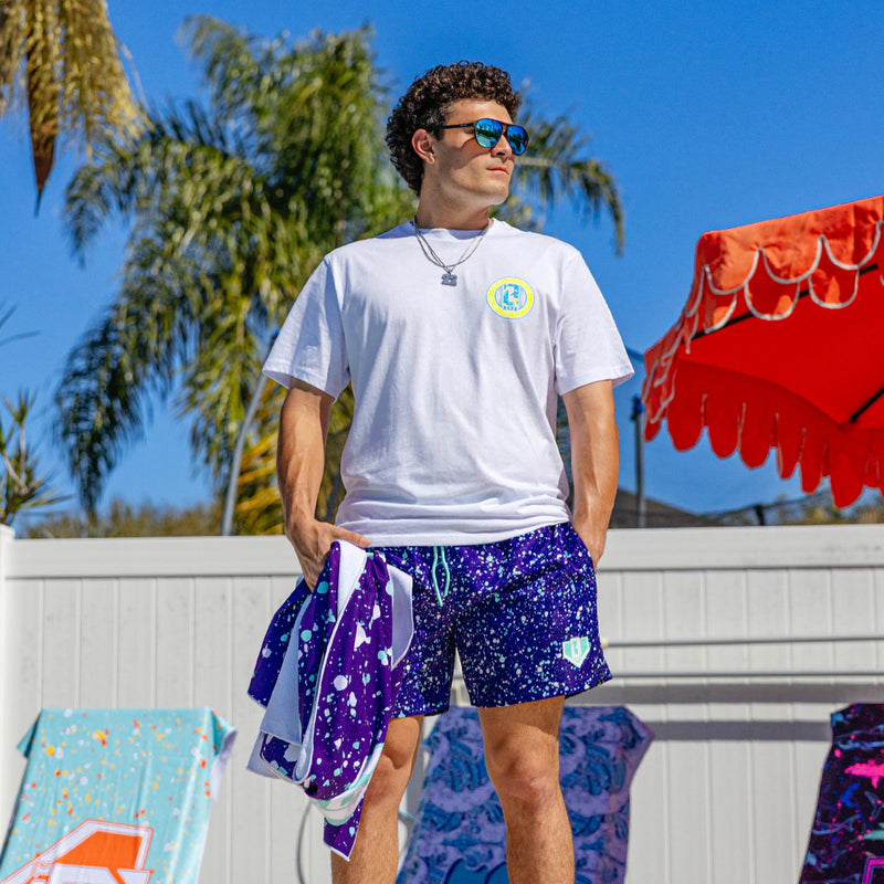 Lifestyle shot of a person wearing BL101 purple splatter swim trunks outdoors with a white tee.