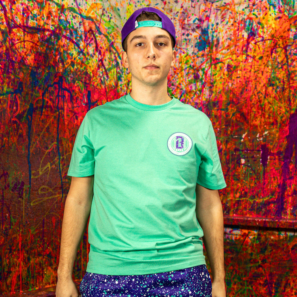 Person wearing a teal t-shirt with a logo and purple cap in front of a colorful abstract wall.