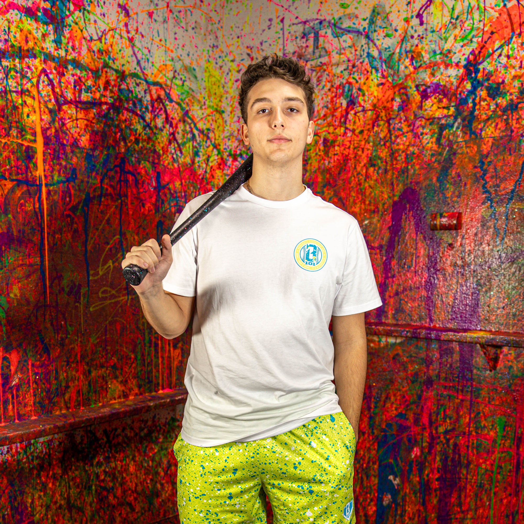 Person wearing a white t-shirt and green splatter shorts, holding a baseball bat against a colorful, abstract wall.