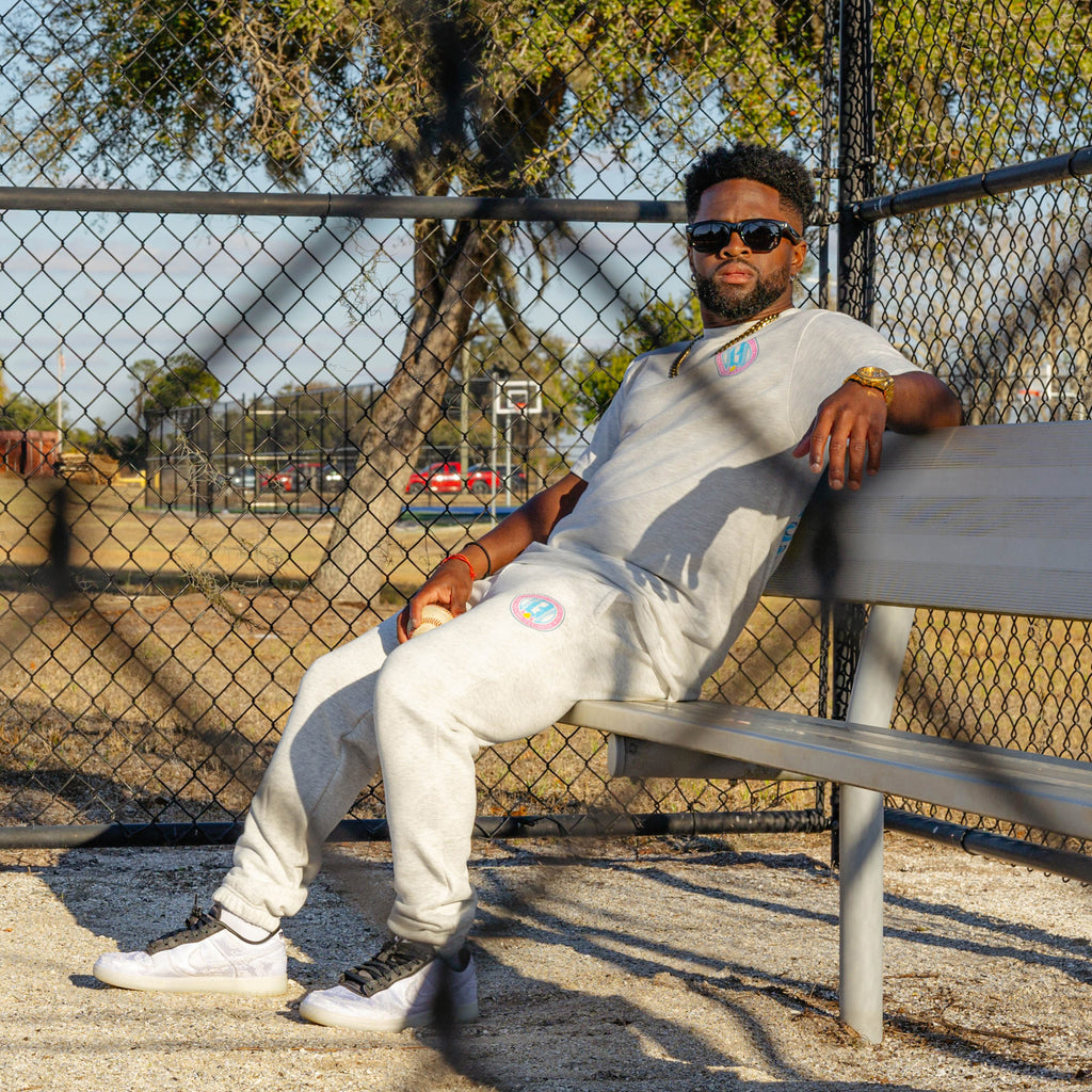 Man sitting on a bench in a park-like setting with a grey t-shirt and grey sweatpants with colorful BL101 logo and trees in the background.