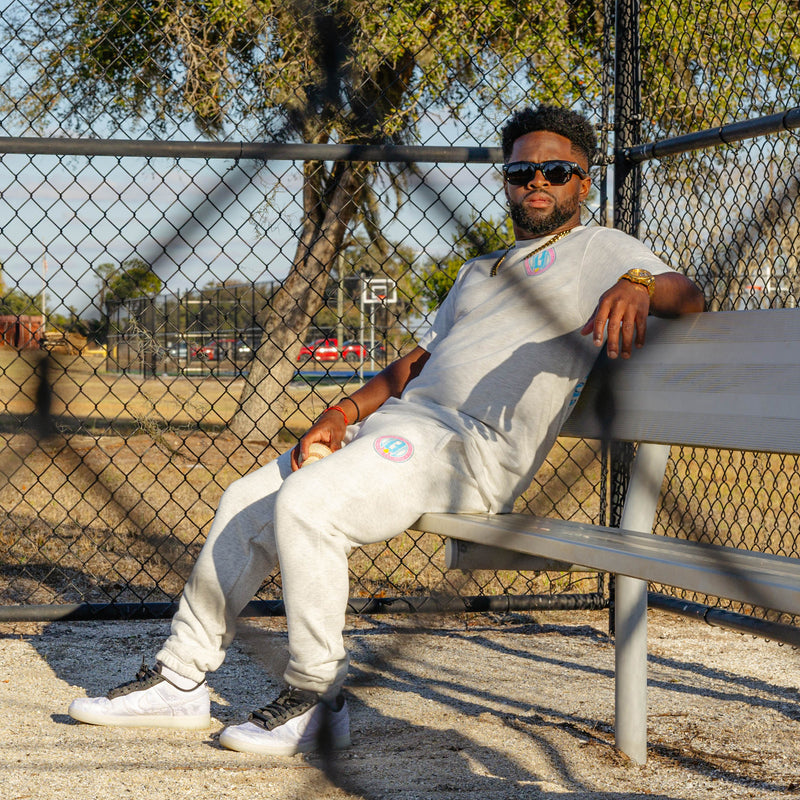 Man sitting on a bench in a park-like setting with a grey t-shirt and grey sweatpants with colorful BL101 logo and trees in the background.