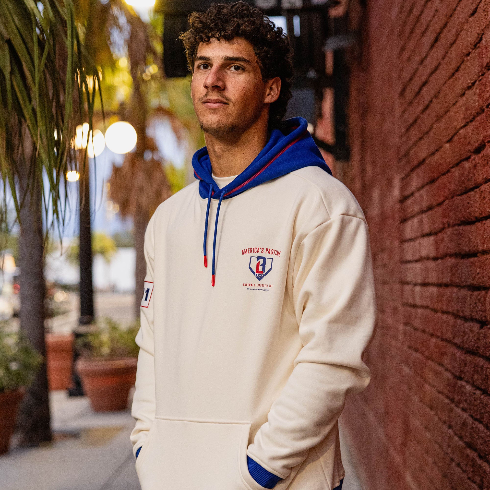 Man leaning against a brick wall wearing a cream stadium hoodie with a royal blue hood