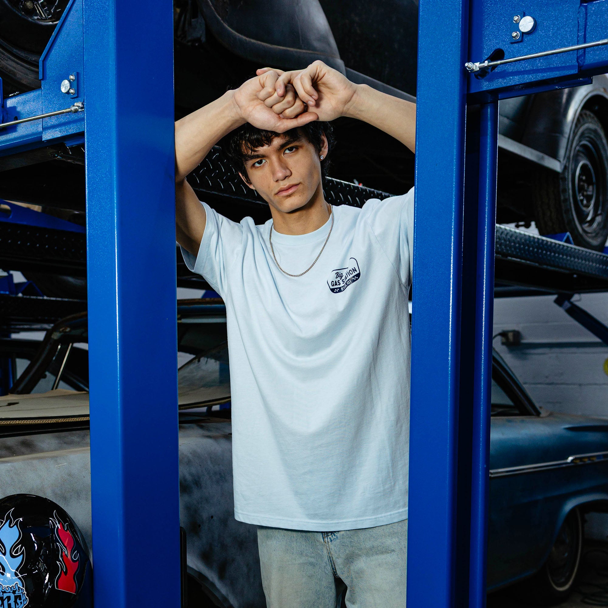 Young man between blue automotive lift columns, arms raised, wearing light blue Big Al's Gas Station tee and light wash jeans.
