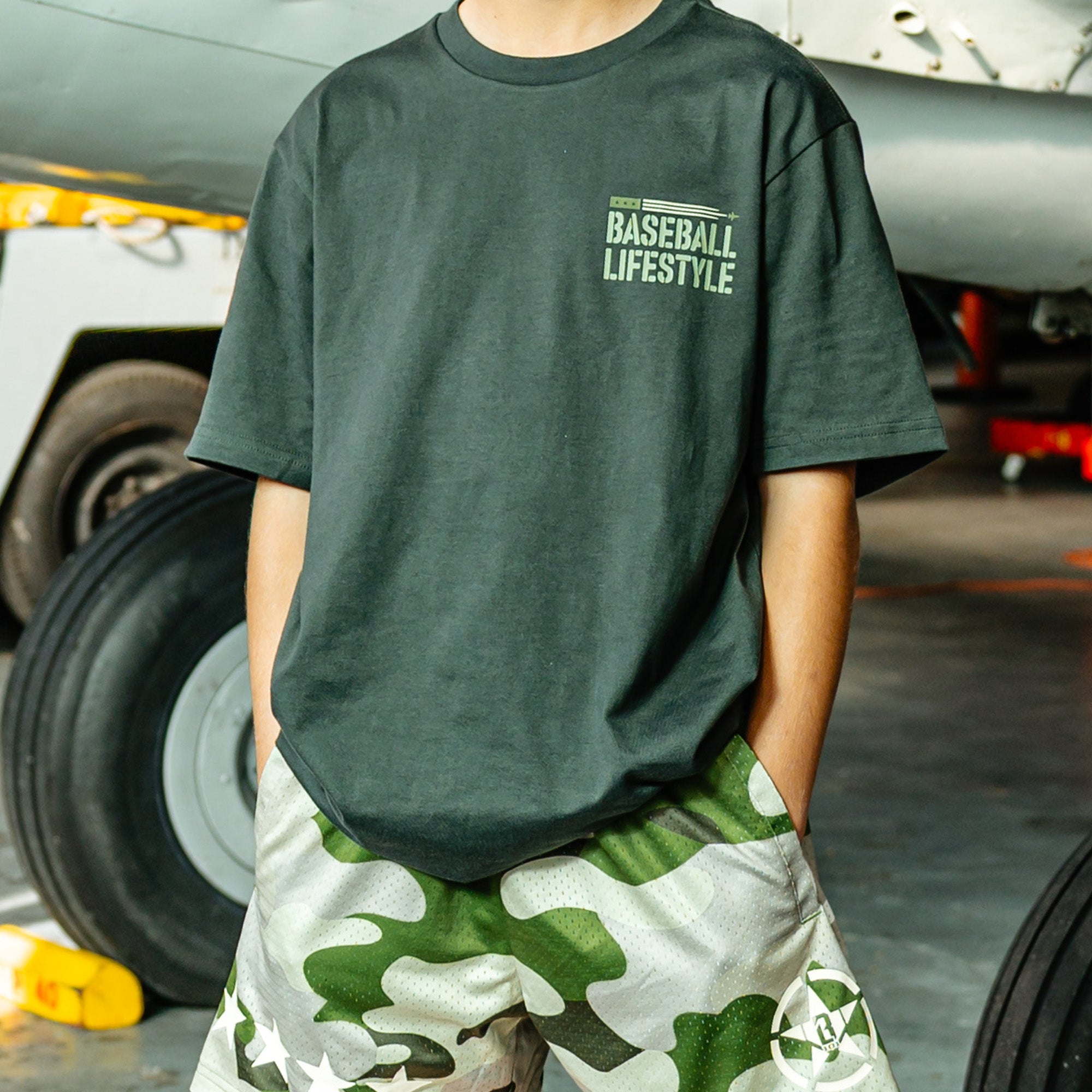 Person wearing a Dark Grey T- shirt with green lettering that says 'BASEBALL LIFESTYLE' in the top right corner