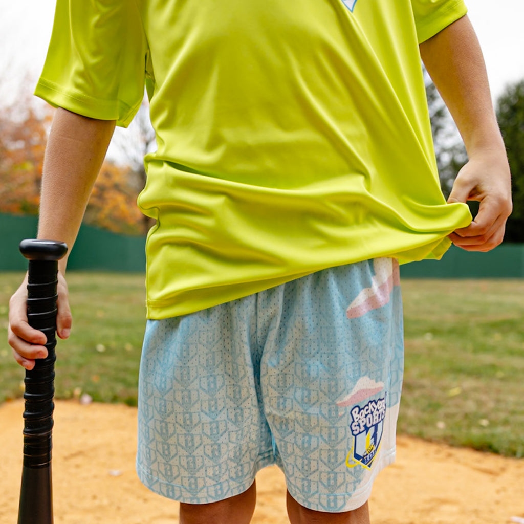 Person wearing a bright yellow shirt and light blue shorts with a 'Backyard Sports BL101' logo, holding a black bat on a baseball field.