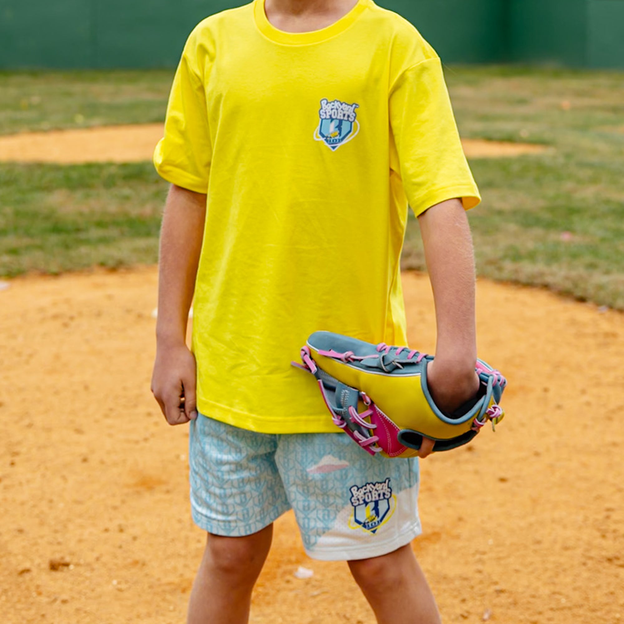 Person wearing a yellow shirt 'BL101 Backyard Sports' and blue shorts with a logo, holding a colorful bag on a dirt field.
