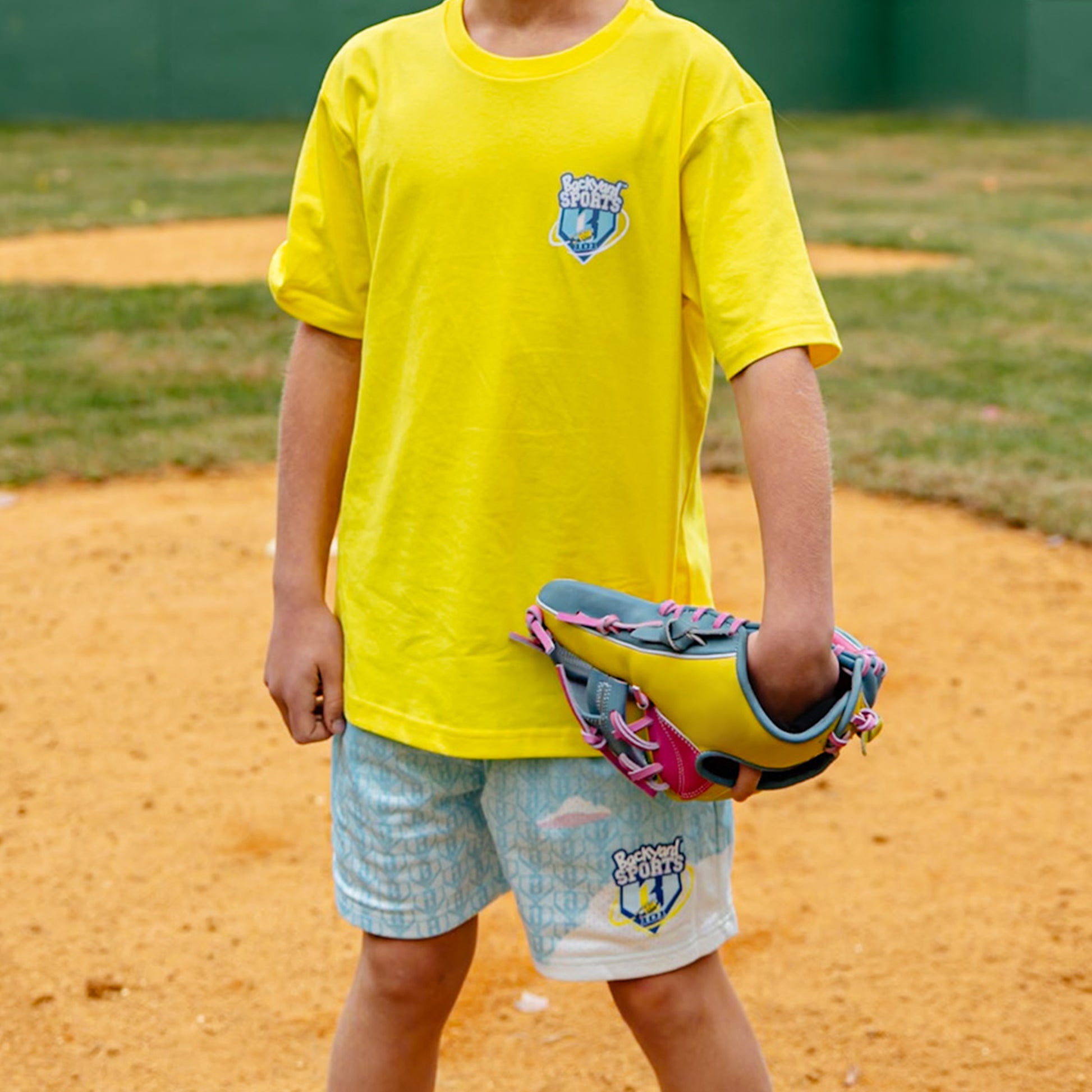 Person wearing a yellow shirt 'BL101 Backyard Sports' and blue shorts with a logo, holding a colorful bag on a dirt field.