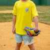 Person wearing a yellow shirt 'BL101 Backyard Sports' and blue shorts with a logo, holding a colorful bag on a dirt field.