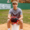 Person sitting on a baseball base with a glove, wearing a gray Pablo Sanchez. shirt and red shorts, on a baseball field.