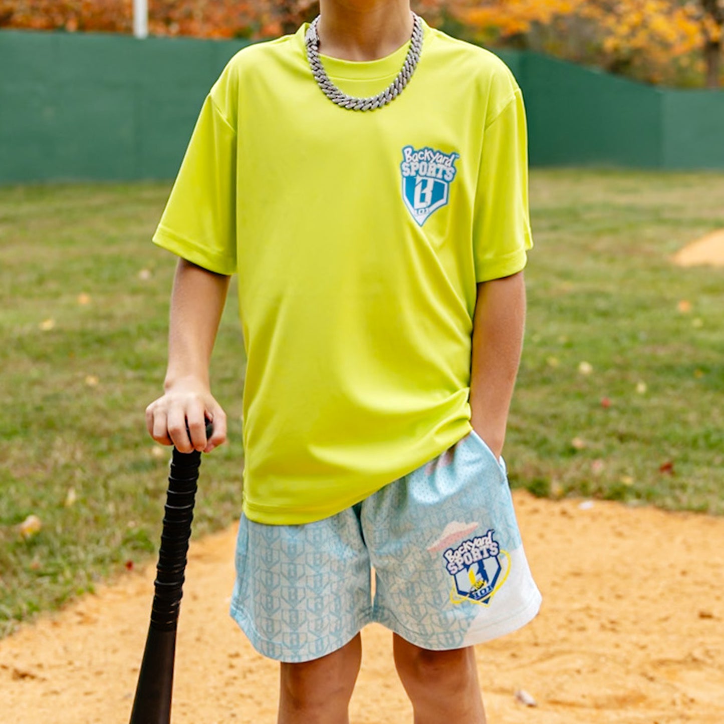 Person wearing a bright yellow sports shirt and light blue shorts with logos, holding a baseball bat on a grassy field.