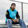 Person wearing a blue, yellow and black varsity jacket with yellow smiley faces, sitting on a bench against a fence.