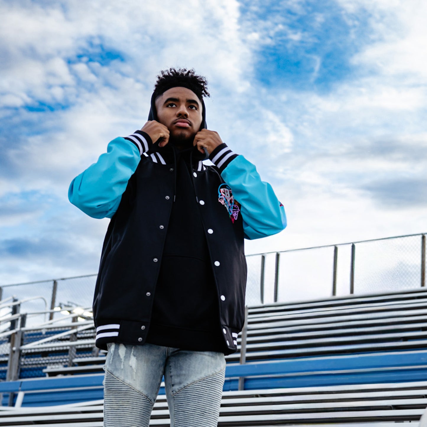 Person wearing a black and blue bomber jacket with a hood, standing against a cloudy sky.