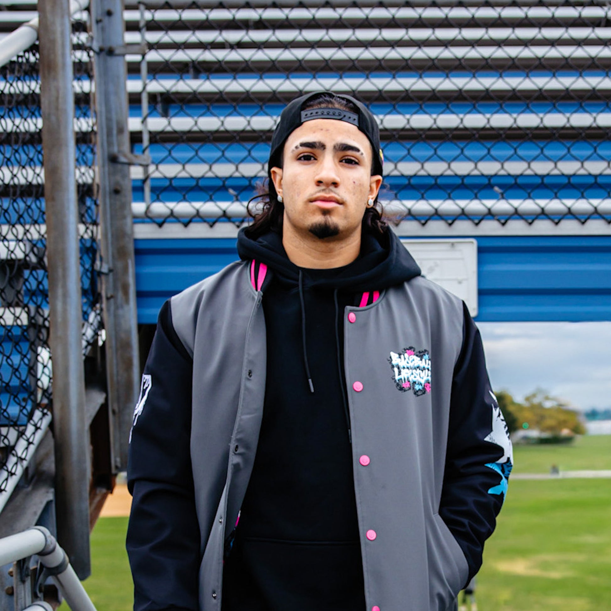 Person wearing a gray and black bomber jacket with a Baseball Lifestyle logo on a sports field background