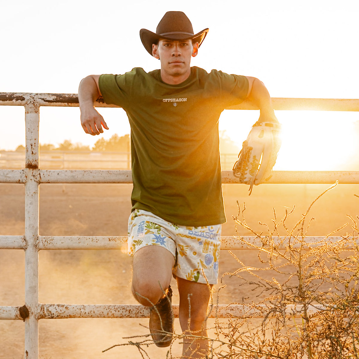 Man wearing a cowboy hat and green 'OFFSEASON' shirt leaning against a fence with a sunset in the background