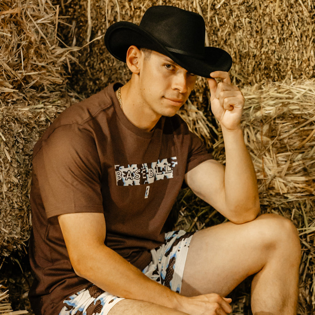 Man wearing a cowboy hat and brown t shit with cow print sitting on hay