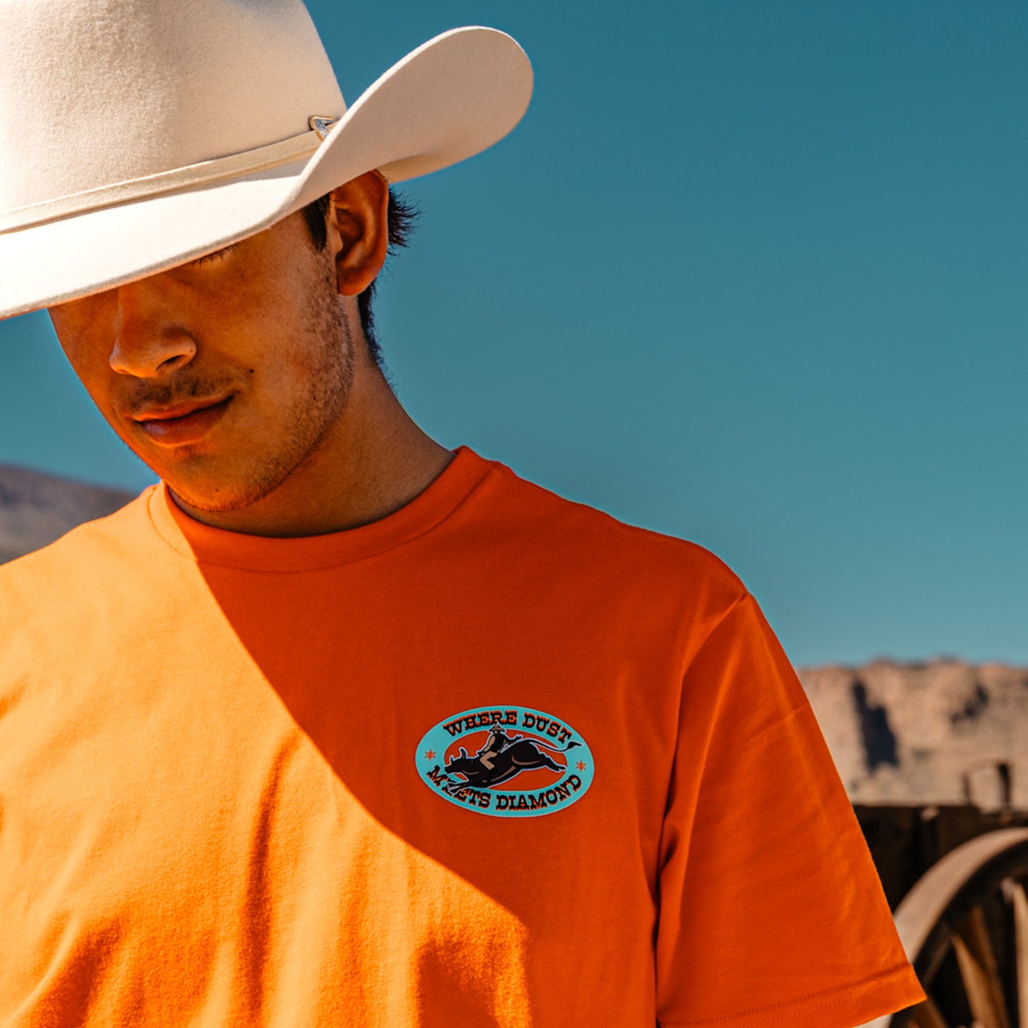 Man wearing an orange t-shirt with a 'WHERE DUST MEETS DIAMOND' logo and a cowboy hat against a clear blue sky.