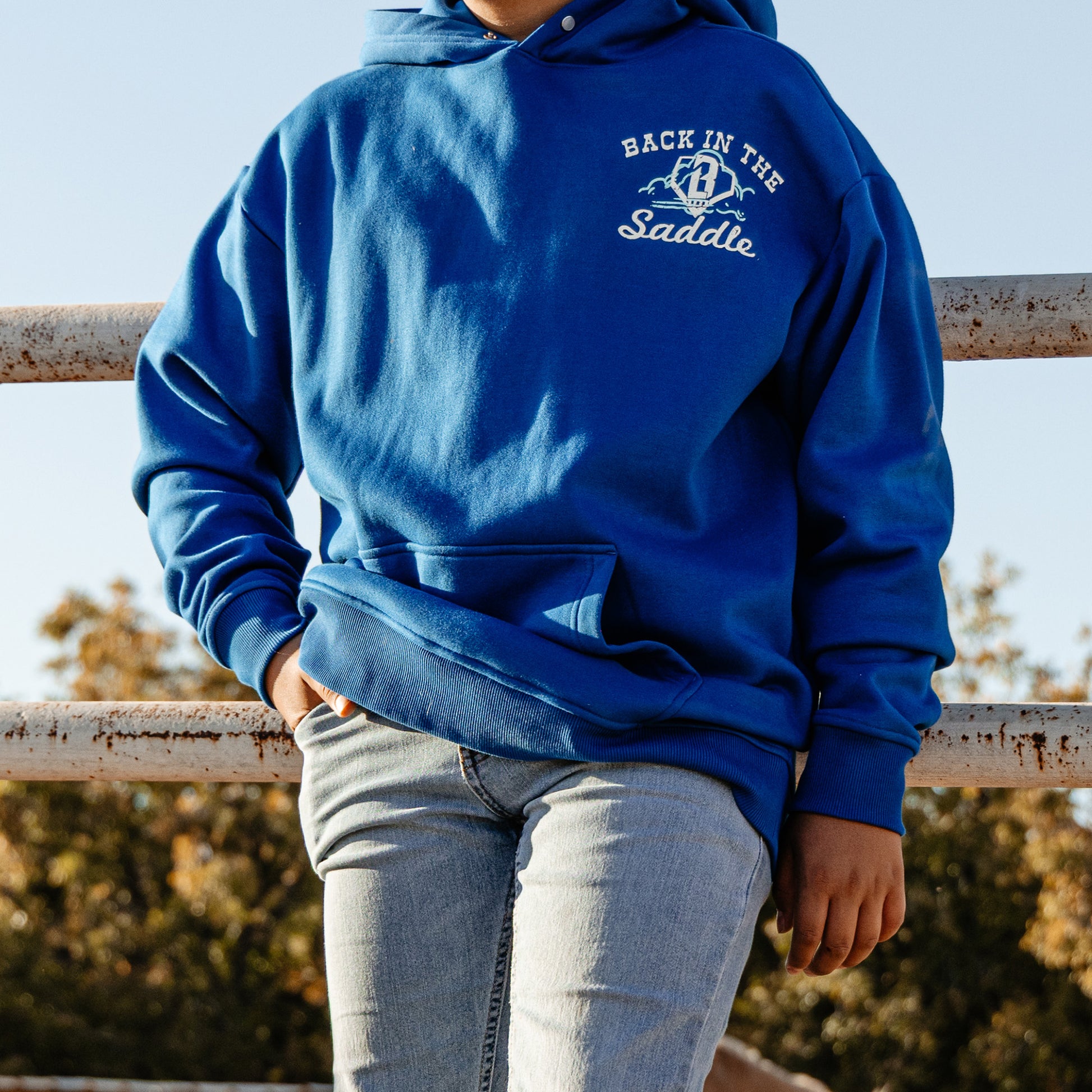 Person wearing a blue hoodie with 'Back in the Saddle' text, standing against a natural background.