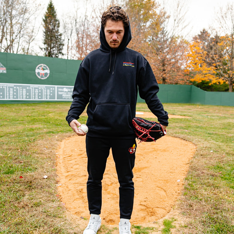 Person in a black hoodie and pants holding a baseball and glove on a baseball field.