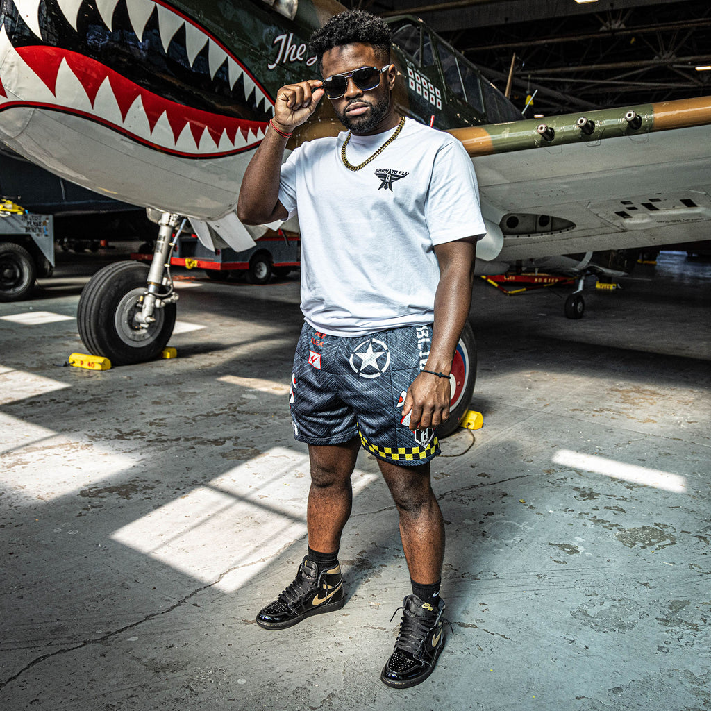 Man wearing a Military-inspired black athletic shorts featuring shark mouth graphics, a white star emblem, black lightening bolts, white BL101 logos on the sides, a green waistband, and yellow checkerboard trim.